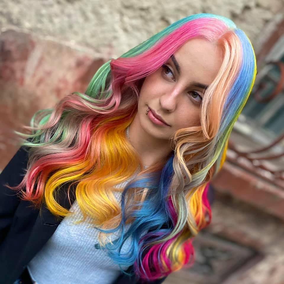 A person with long, wavy hair dyed in vibrant rainbow colors including pink, green, blue, yellow, and orange, stands outside near a textured wall. They are wearing a lip piercing, a black jacket over a white top, and are looking slightly to the side.