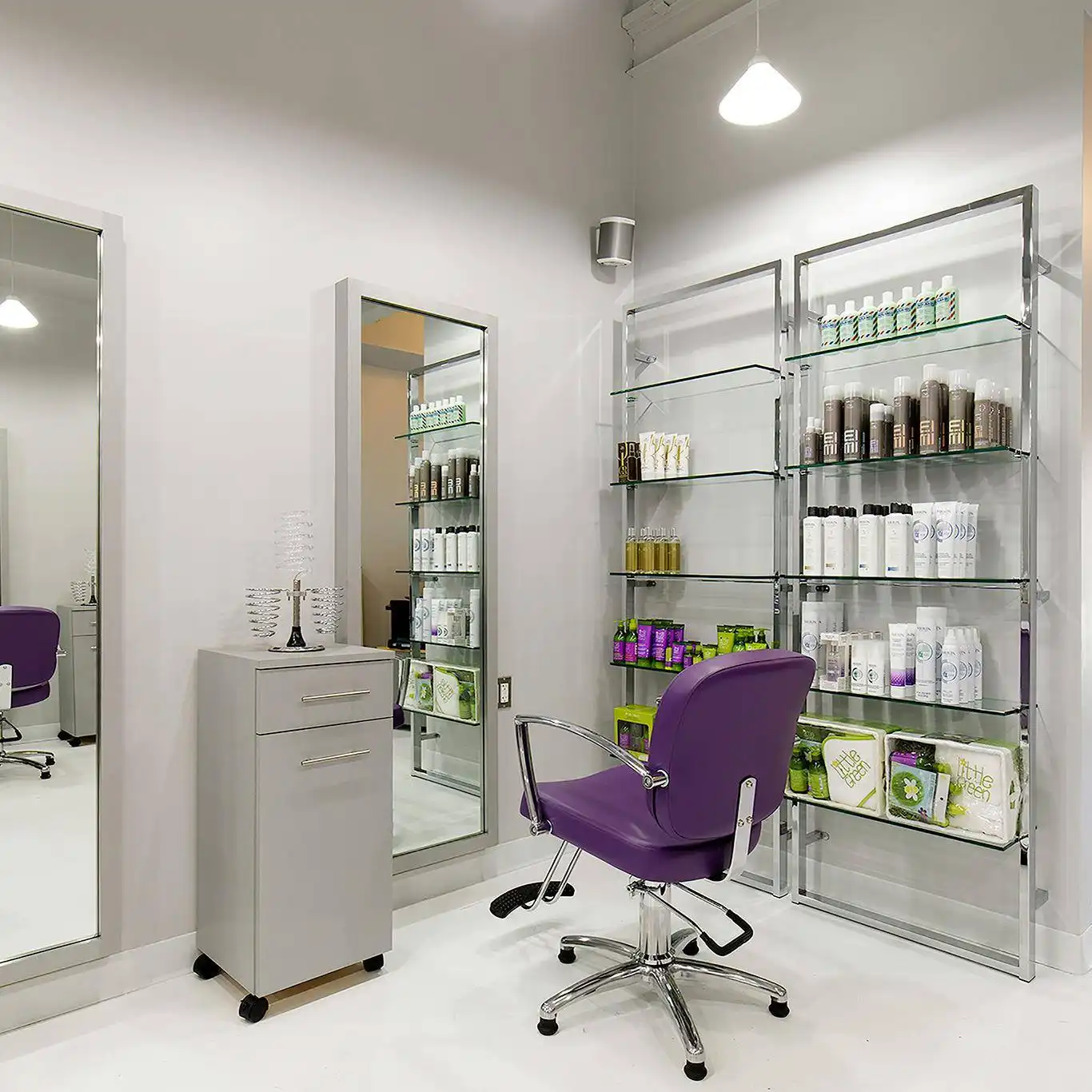 A modern salon interior featuring a styling station with a purple chair and mirror. Nearby, a tall cabinet holds various hair care products neatly arranged on glass shelves. A white wall and ceiling with overhead lighting complete the clean, minimalist look.