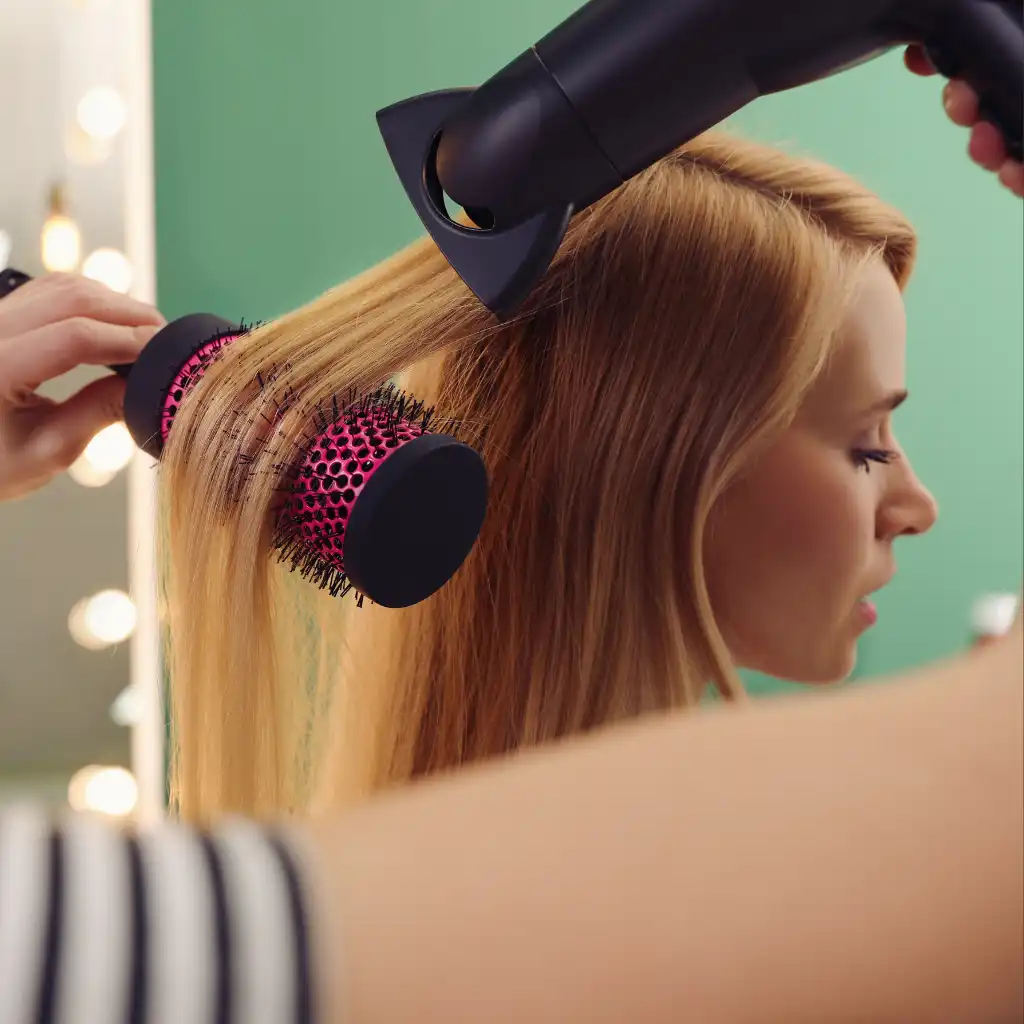 Close-up of a woman with long blonde hair being blow-dried and styled with a round brush in a salon. The background features a green wall and blurred vanity lights. The stylist’s hand holds the brush while the hairdryer is directed at the hair.