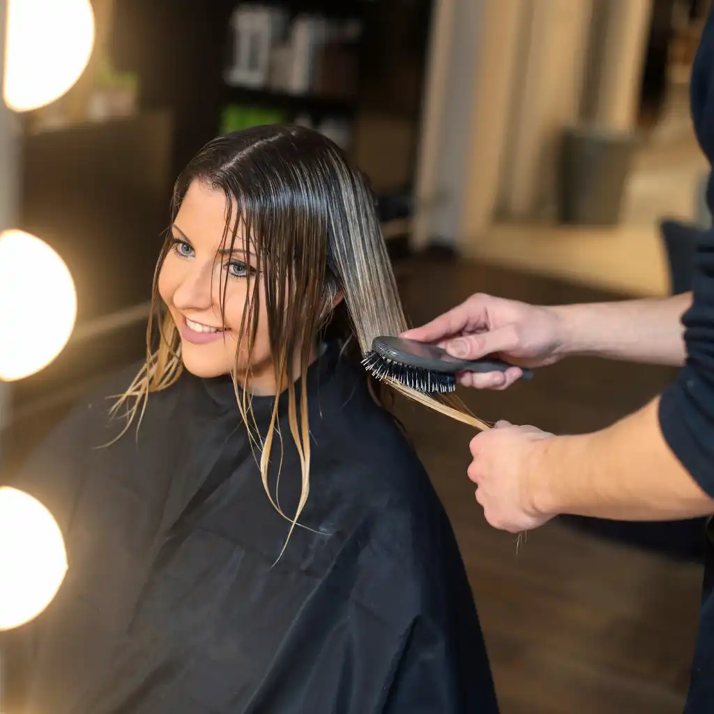 A woman with wet hair is sitting in a salon chair, draped with a black cape. She is smiling while a stylist, whose face is not visible, brushes her hair. There are illuminated vanity lights in the foreground, and shelves with products in the background.