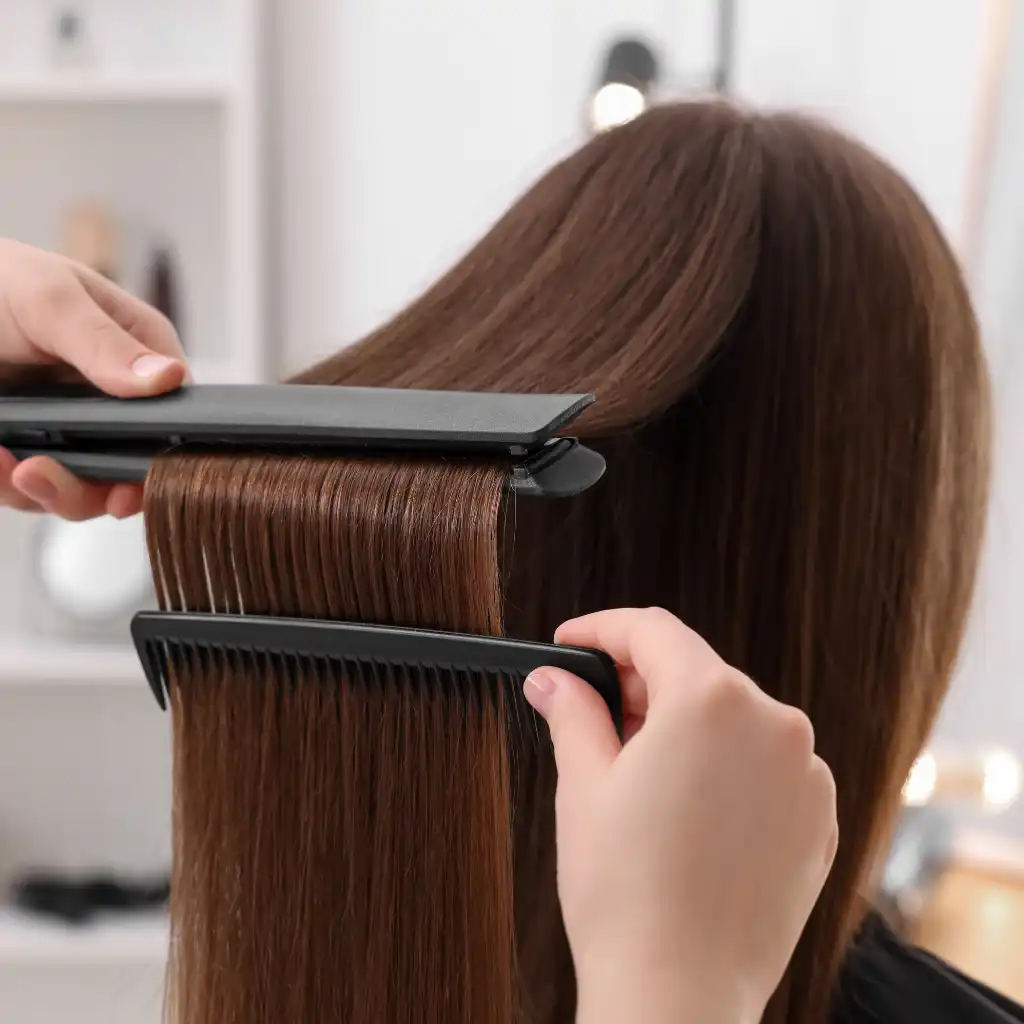 A person is getting their long, straight brown hair styled with Hot Tools Styling. One hand uses a flat iron to straighten the hair, while the other holds a black comb to ensure each section is smooth. The background shows a blurred salon setting with shelves and lights.