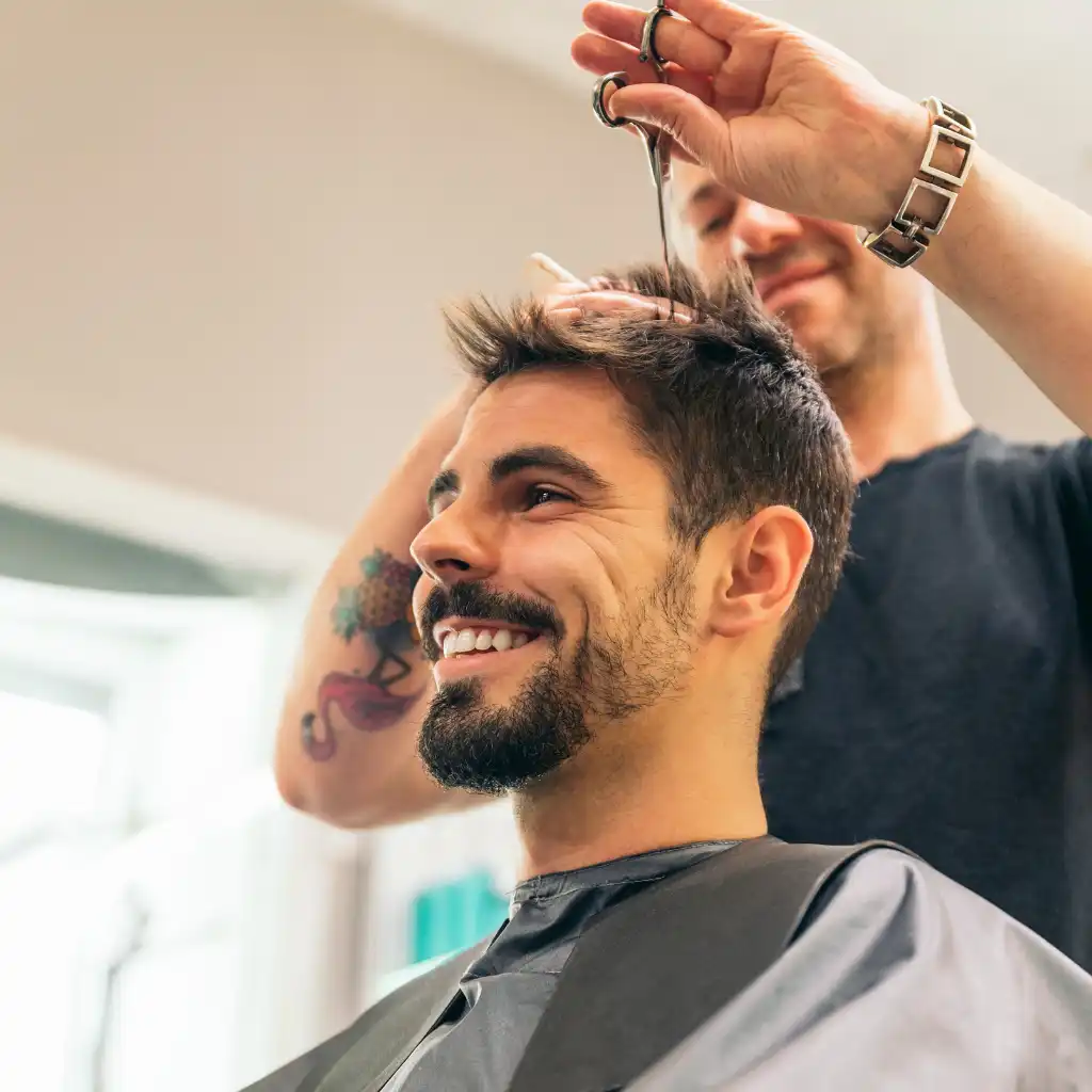 A smiling man with a beard is getting a haircut. A barber, partially visible, is cutting the man's hair with scissors. The man is wearing a black cape and the background shows a blurred interior with natural light coming through a window.