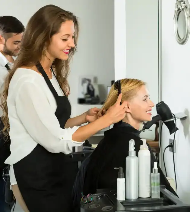 A hairstylist with long brown hair and wearing a black apron is smiling while brushing the hair of a woman with blonde hair, who is seated in a salon chair and also smiling. Various hair products and tools are visible on the table in front of them.
