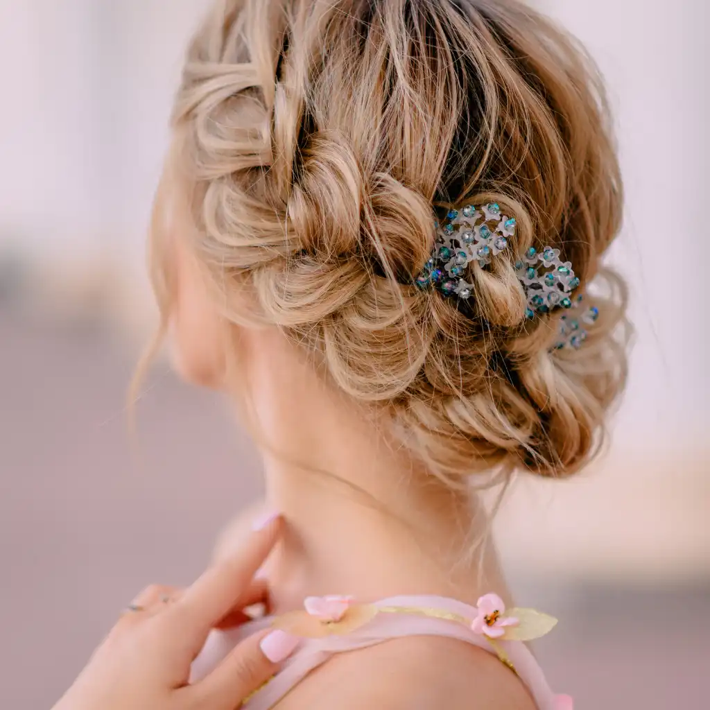 A woman's blonde hair is styled in an elegant updo with intricate braids. A decorative hairpin adorned with blue and green jewels secures part of the braid. She is wearing a light pink top with floral accents and has her fingers gently touching her neck.