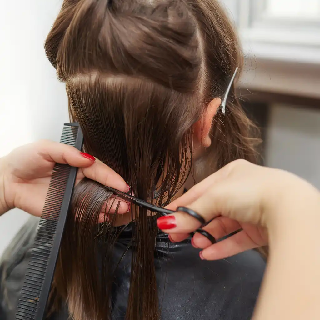 A young woman is getting a haircut. The hairdresser is holding a section of the person's long, brown hair with a comb in one hand and cutting it with scissors in the other hand. The person's hair is partially pinned up with a hair clip. The setting appears to be a salon.